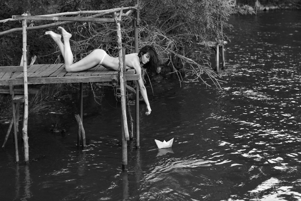 nude woman reaching for a paper ship in the water below