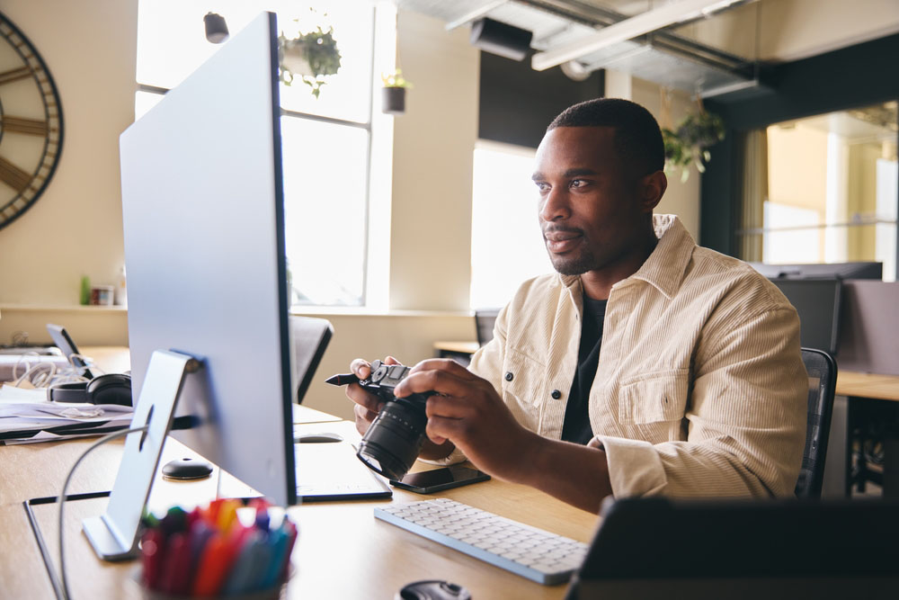 Young Black Professional Photographer Sitting At Desk Working On Computer Holding Camera Editing Pictures