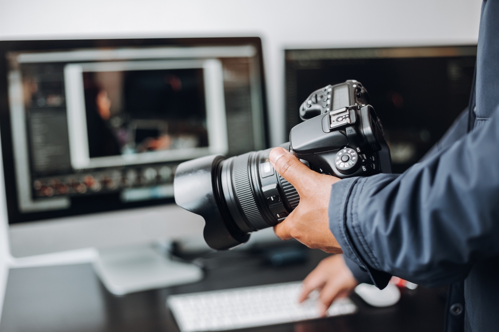 photographer's hand on their camera and a computer on a desk