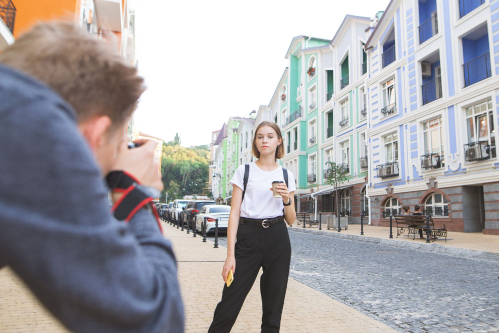 Professional photographer photographs a girl on the street. Girl model with a glass of coffee in her hands poses a photographer on the street of the town. Photosession concept