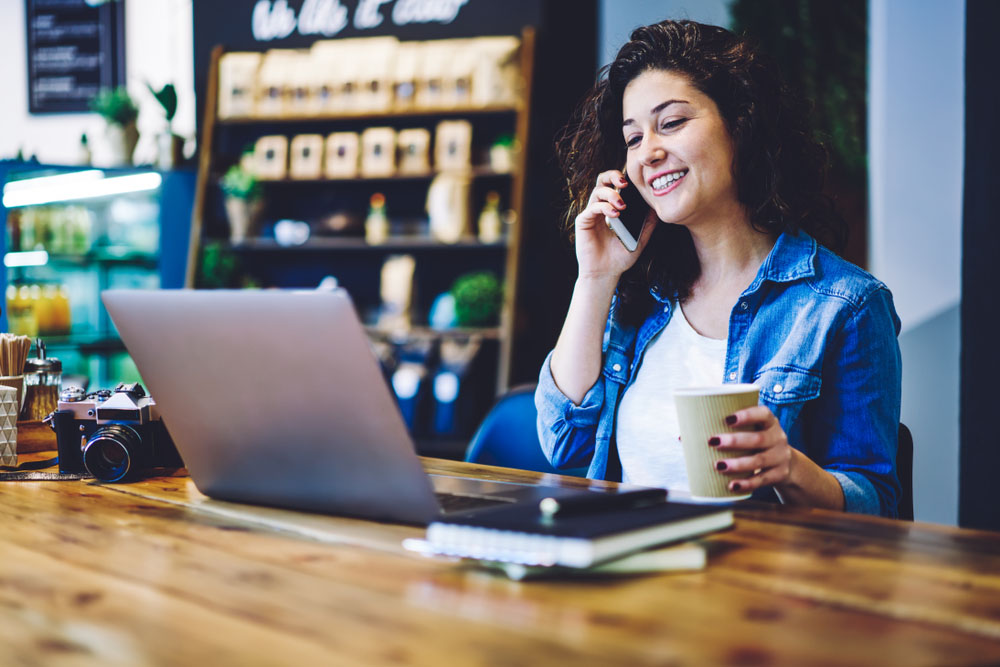 Cheerful woman entrepreneur calling to colleague during coffee break in coworking space using 4g internet on smartphone gadget, happy female photographer watching webinar with useful information